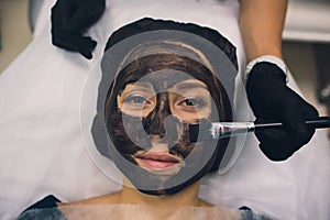 The cosmetologist in pink gloves with a brush applies a carbon mask for peeling on the face of a young girl in a