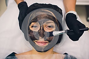 The cosmetologist in pink gloves with a brush applies a carbon mask for peeling on the face of a young girl in a