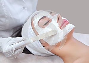 The cosmetologist for the procedure of cleansing and moisturizing the skin, applying a mask with stick to the face of a young woma