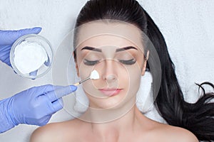 The cosmetologist for the procedure of cleansing and moisturizing the skin, applying a mask to the face of a young woman