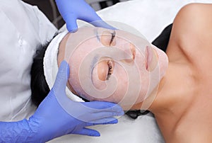 The cosmetologist for procedure of cleansing and moisturizing the skin, applying a sheet mask to the face of a young woman in beau