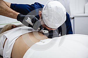 A cosmetologist in a white cap and black gloves examines a mole on the patient& x27;s back using a dermatoscope