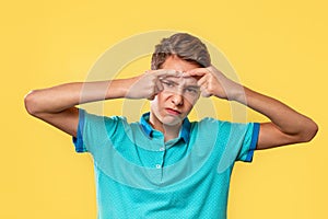 Cosmetology, Dermatology and acne. Caucasian teenager in a blue t-shirt, painfully squeezes out a pimple on his forehead. Yellow