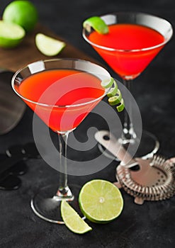 Cosmopolitan cocktail in classic crystal glasses with lime peel and fresh limes with strainer on black background