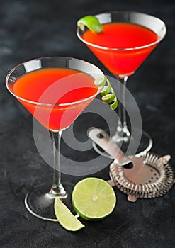 Cosmopolitan cocktail in classic crystal glasses with lime peel and fresh limes with strainer on black background