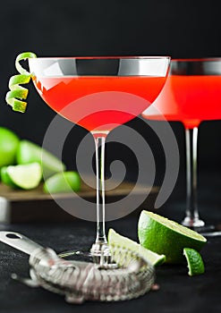 Cosmopolitan cocktail in modern crystal glasses with lime peel and fresh limes with strainer on black background