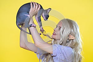 Cosplayer elf young female in blue dress and Sphinx kitten look at each other. Yellow background