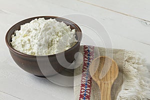 Cottage cheese in a brown ceramic bowl with authentic flax embroidered napkin and a wooden spoon on a white wooden background. C