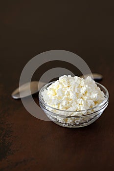 Cottage cheese crumbly in a glass bowl and spoon on a wooden old brown tabletop