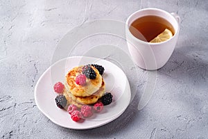 Cottage cheese pancakes, curd fritters dessert with raspberry and blackberry berries in plate near to hot tea cup with lemon slice