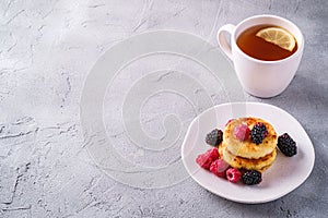 Cottage cheese pancakes, curd fritters dessert with raspberry and blackberry berries in plate near to hot tea cup with lemon slice