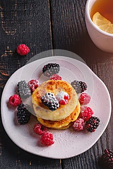 Cottage cheese pancakes, curd fritters dessert with raspberry and blackberry berries in plate near to hot tea cup with lemon slice