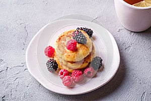 Cottage cheese pancakes, curd fritters dessert with raspberry and blackberry berries in plate near to hot tea cup with lemon slice