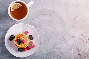 Cottage cheese pancakes, curd fritters dessert with raspberry and blackberry berries in plate near to hot tea cup with lemon slice