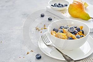 Cottage cheese with pieces of pears, nuts and blueberries in a white cup on a light-gray background. Delicious and