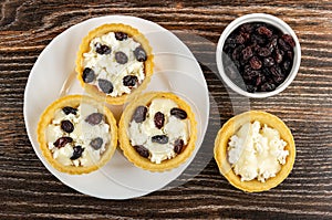 Cottage cheese with raisin and condensed milk in tartlets in plate, bowl with dried grape on wooden table. Top view