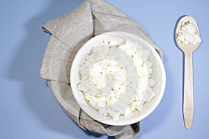 Cottage cheese in white bowl on blue table with spoon and napkin. Top view.