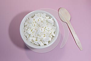 Cottage cheese in white bowl on pink table with spoon. Top view.