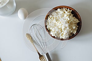 Cottage cheese in a wooden cup golden fork and spoon .kitchen whisk and egg on a white table