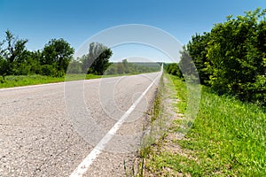 Country asphalt road in summer with green grass and trees on the roadsides against the blue sky. Sunny day background