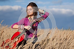 Country girl fixing her hair and holding an acoustic guitar in field against blue cloudy sky background