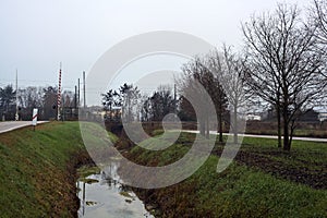 Country road bordered by a stream of water that leads to a railroad crossing on a cloudy day