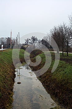 Country road bordered by a stream of water that leads to a railroad crossing on a cloudy day