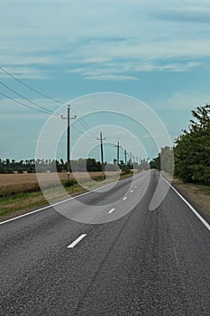 A country road with electric poles. Electric poles along a village road. Nearby a field of wheat. Blue sky with clouds