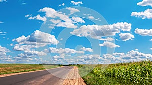 A country road goes over the horizon under clouds in a bright blue sky between a freshly mown field and a field of corn.
