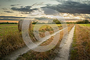 Country road and hay bale in the field, evening view with clouds