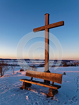 Wooden cross and bench on hill in snow-covered countryside by sunset light