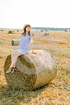 Countryside landscape of farm wheat field at summer sunset. Pretty young happy Caucasian woman in straw hat and blue