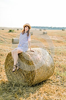 Countryside landscape of farm wheat field at summer sunset. Pretty young happy Caucasian woman in straw hat and blue