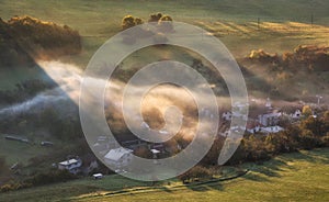 Countryside rural landscape with village over sunbeam in misty morning