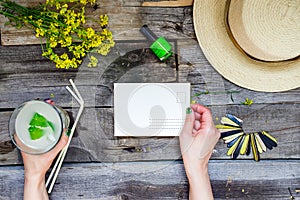 Countryside travel and vacation background. Female hands holding postcard surrounded with glass of lemonade, straw hat, wildflower