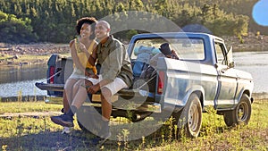 Couple With Backpacks In Pick Up Truck On Road Trip By Lake Drinking Beer