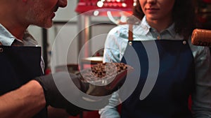 Couple of baristas in uniform checking the quality of roasted coffee beans standing near the roaster machine.