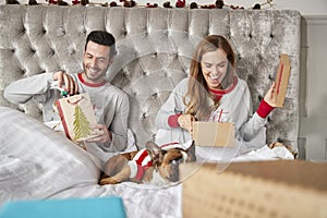 Couple In Bed At Home With Dog Dressed In Santa Costume Opening Gifts On Christmas Day