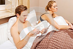 Couple in bed, man using tablet woman sitting bored