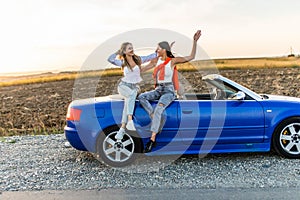 Couple of best women friends having fun together smiling while sitting on convertible car on sunset