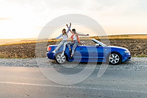 Couple of best women friends having fun together smiling while sitting on convertible car on sunset