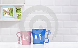 Couple blue and pink coffee mugs on table with blurred decorative shelf on white tile wall