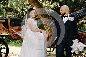 A couple of the bride and groom are standing near the carriage in nature in retro style