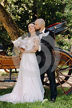 A couple of the bride and groom are standing near the carriage in nature in retro style