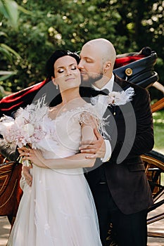 A couple of the bride and groom are standing near the carriage in nature in retro style