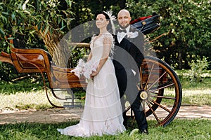 A couple of the bride and groom are standing near the carriage in nature in retro style