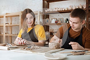 Couple in casual clothes and aprons making ceramic pot on pottery at table in workshop.