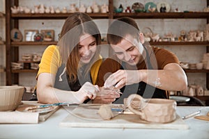 Couple in casual clothes and aprons making ceramic pot on pottery at table in workshop.