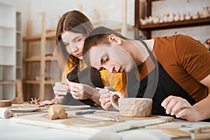 Couple in casual clothes and aprons making ceramic pot on pottery at table in workshop.