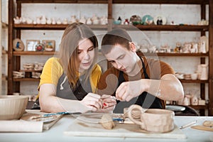 Couple in casual clothes and aprons making ceramic pot on pottery at table in workshop.